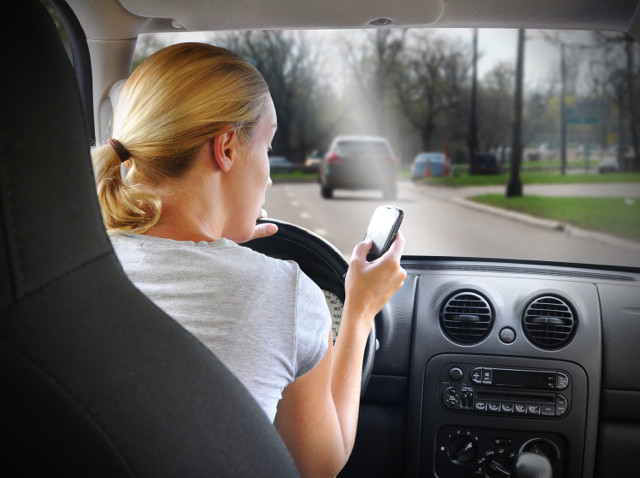 A young woman is on the cell phone textign and driving with a road in the windshield for an danger or distracted driving concept.